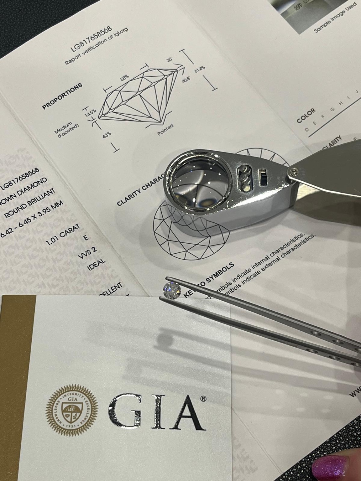 A loose diamond held in tweezers beside a jeweller’s loupe and diamond grading reports